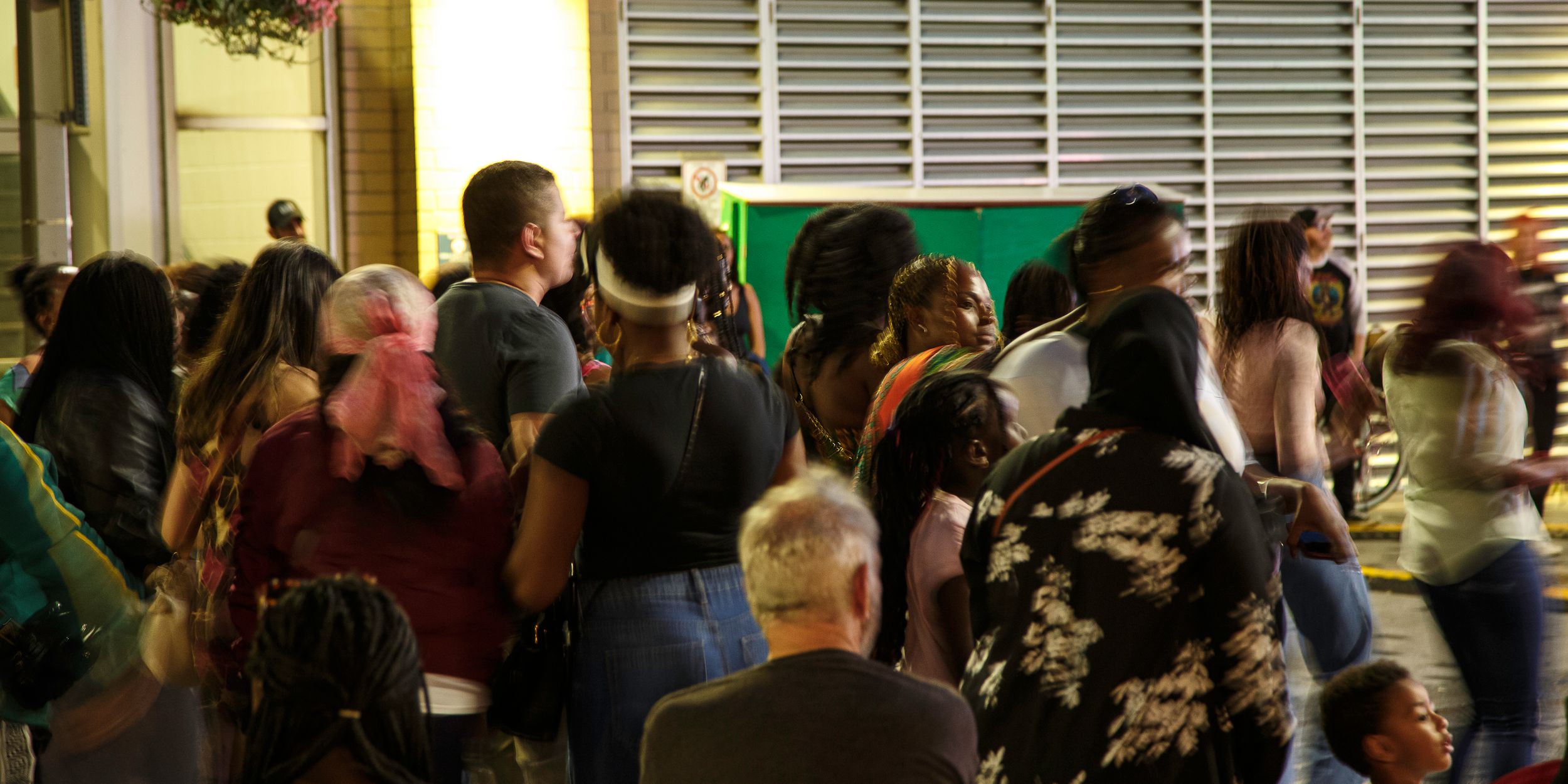 Crowd of people standing in line outdoors at night in front of a building with a lit entrance and louvered metal shutters.