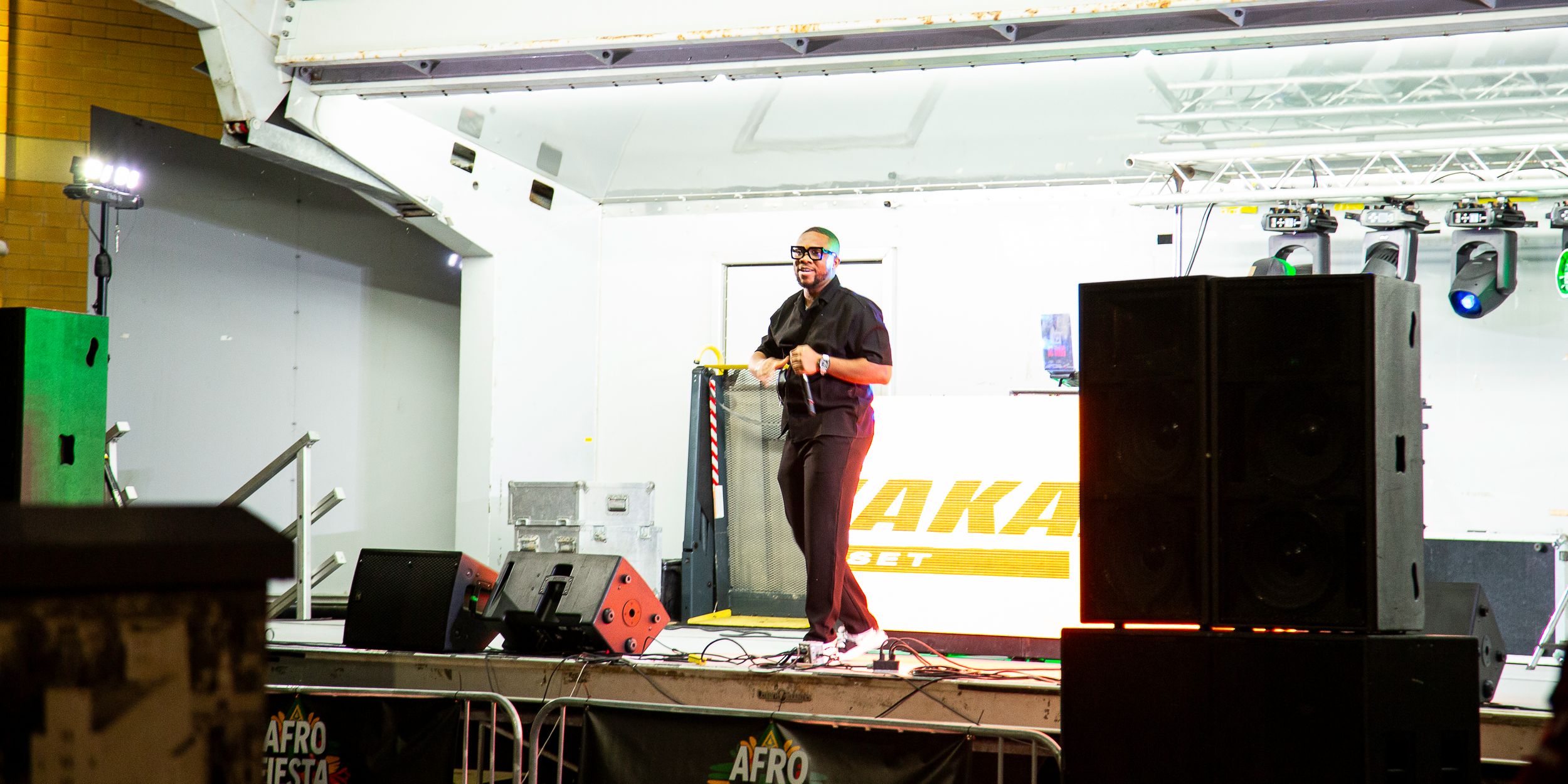 Performer in a cap and dark clothing stands on an outdoor stage beside large black speakers, with a banner and stage lights behind.