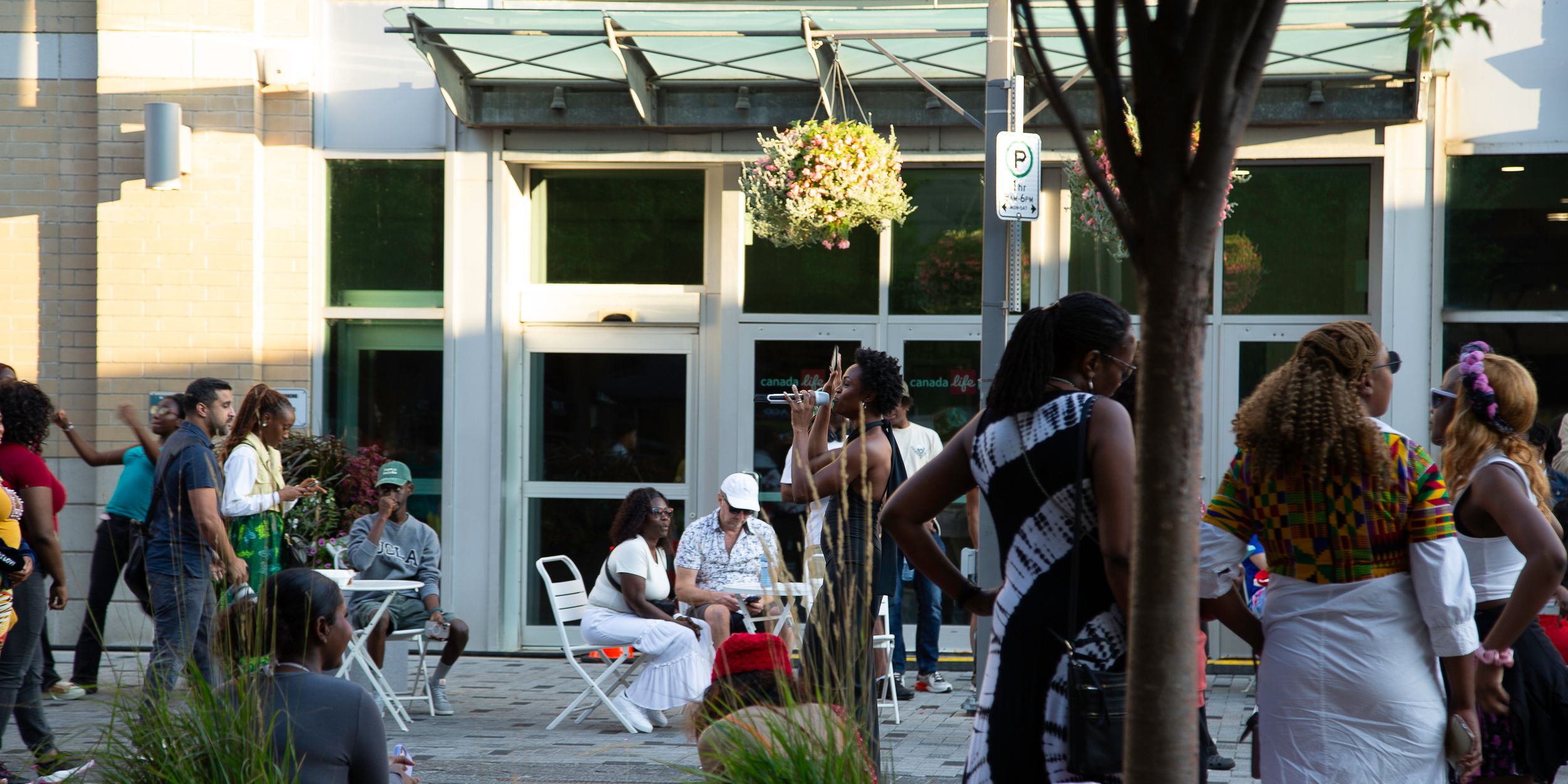 Crowd of people gathering on a sunny urban plaza with outdoor seating, a hanging flower basket, and a modern glass storefront.