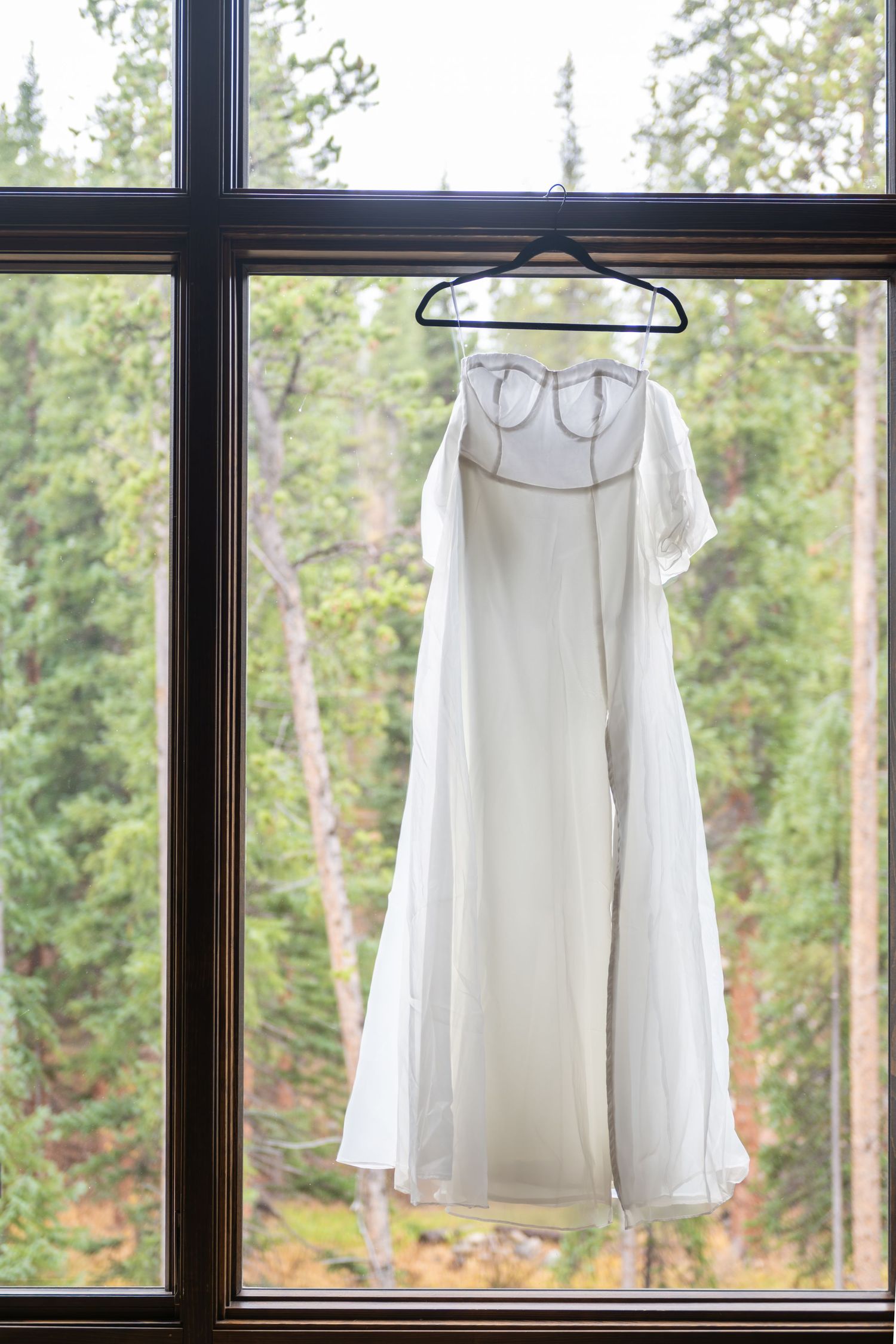 A white wedding dress hangs on a hanger against a window with evergreen trees visible in the background.