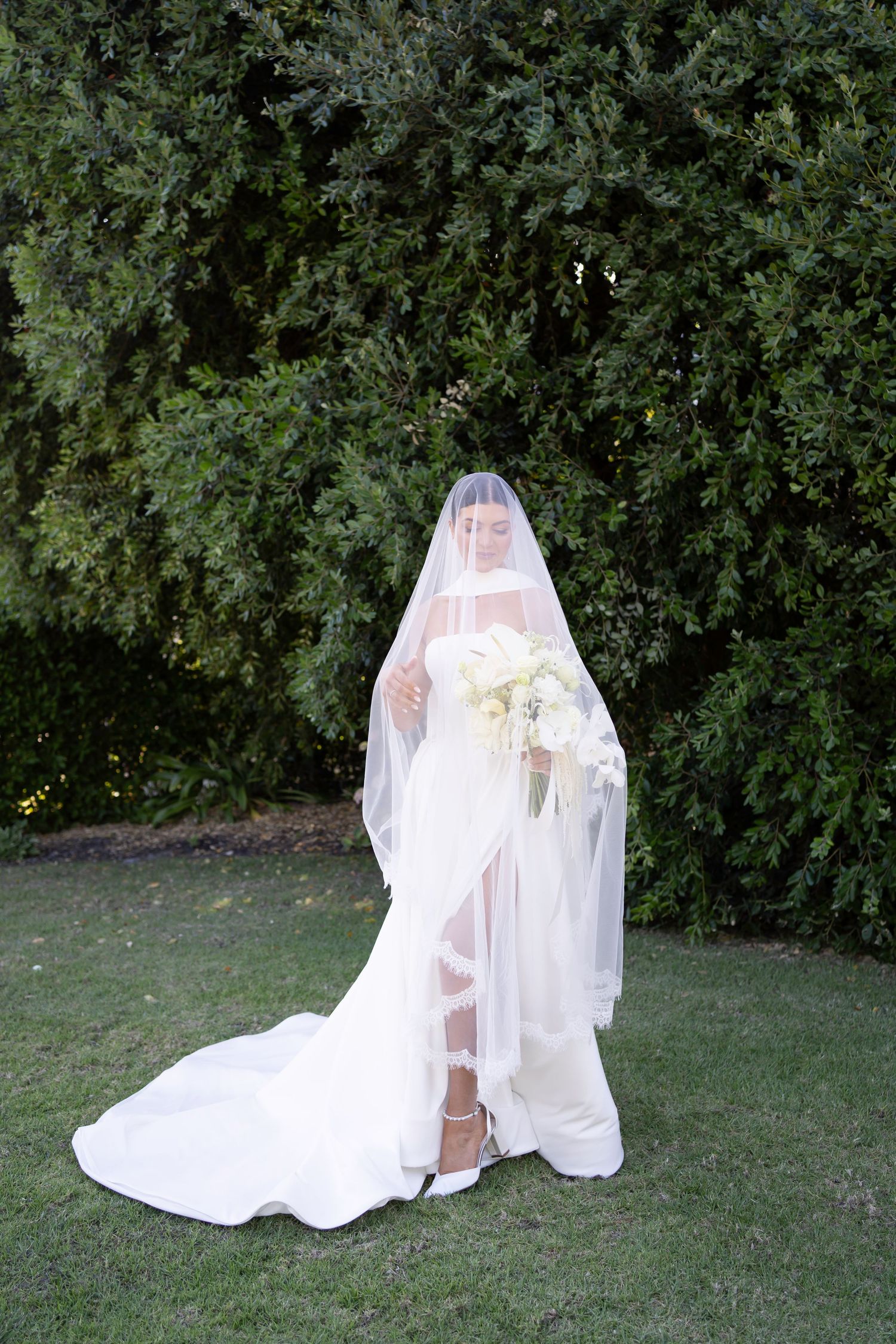 A bride in a flowing white wedding gown and long veil stands on a lawn with lush green foliage in the background.