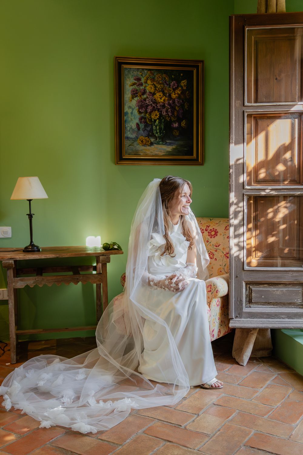 A bride in a flowing white gown sits gracefully on terracotta tiles near a window in a room with sage green walls.