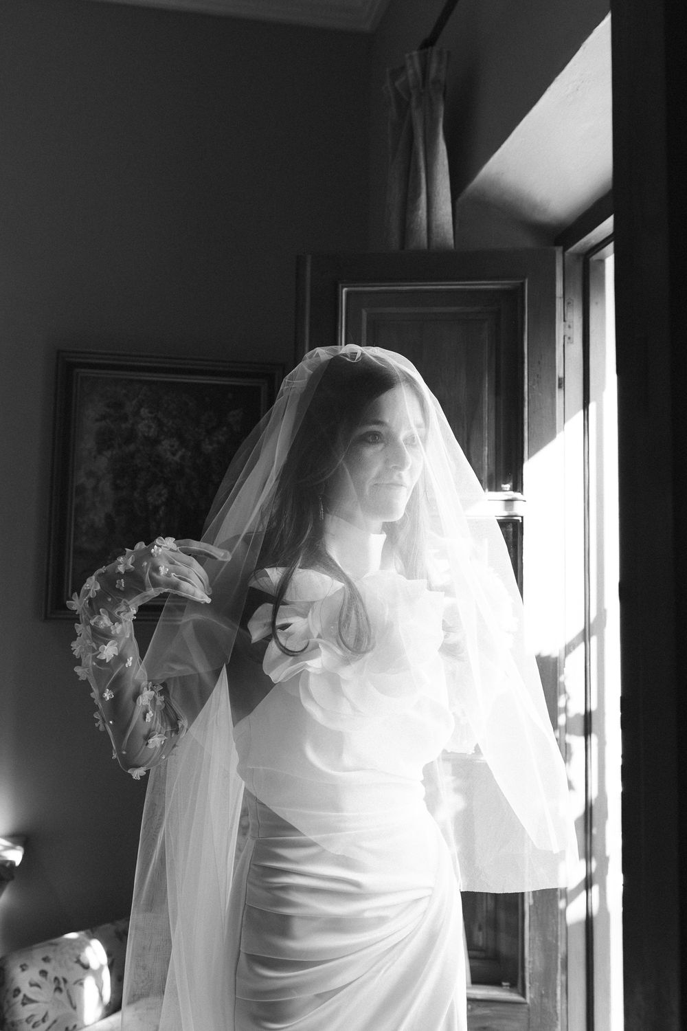 Dramatic window lighting illuminates flowing white fabric and ruffles in an elegant bridal preparation scene.