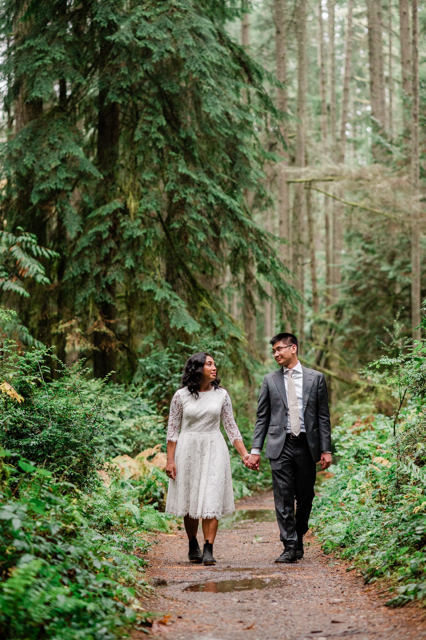 A couple walks hand in hand on the Grand Forest West trail surrounded by tall evergreen trees and lush ferns.