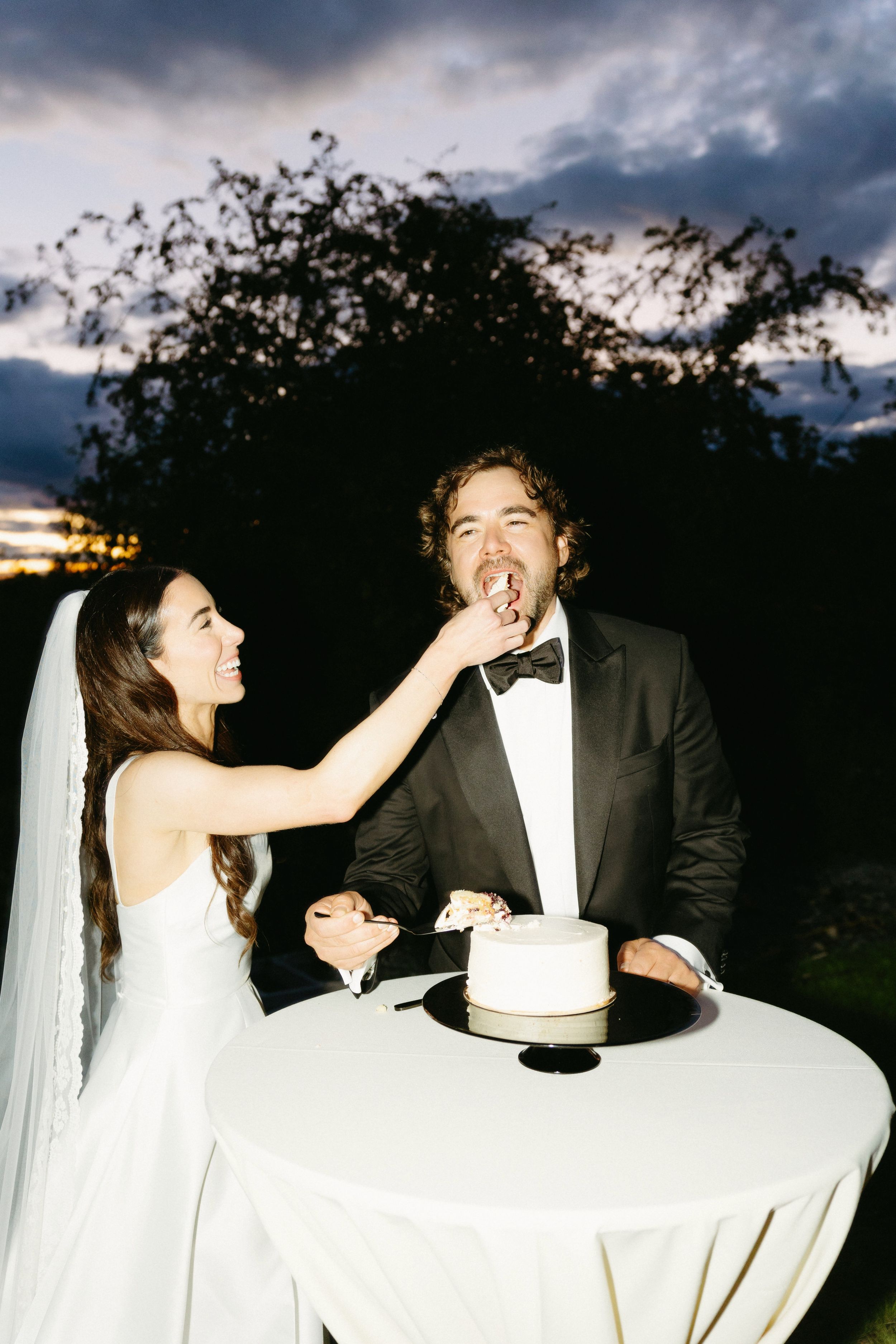 Playful cake cutting moment at an outdoor wedding reception at dusk.