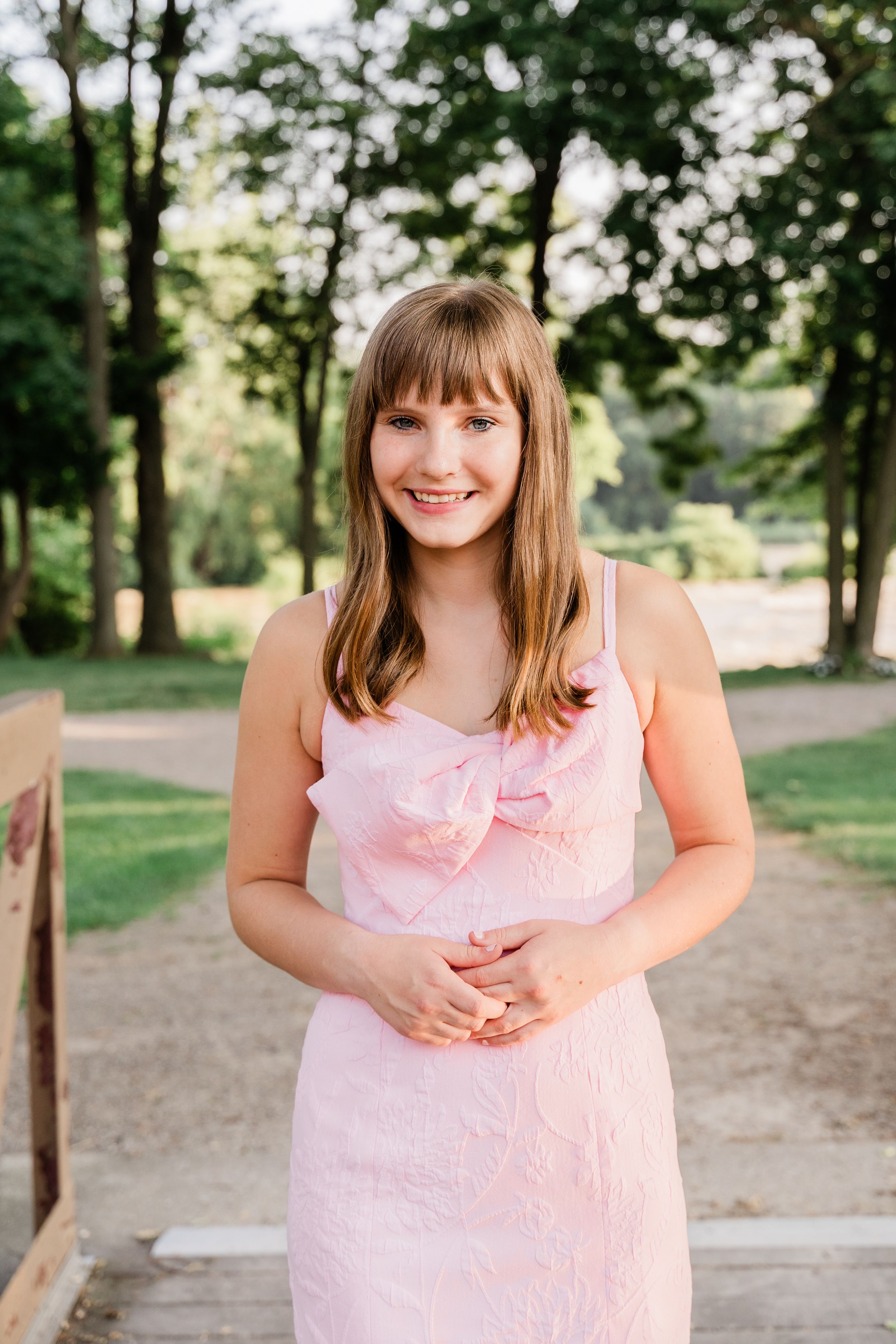 Someone wearing a light pink dress stands on a wooden path surrounded by trees in a park setting on a sunny day.