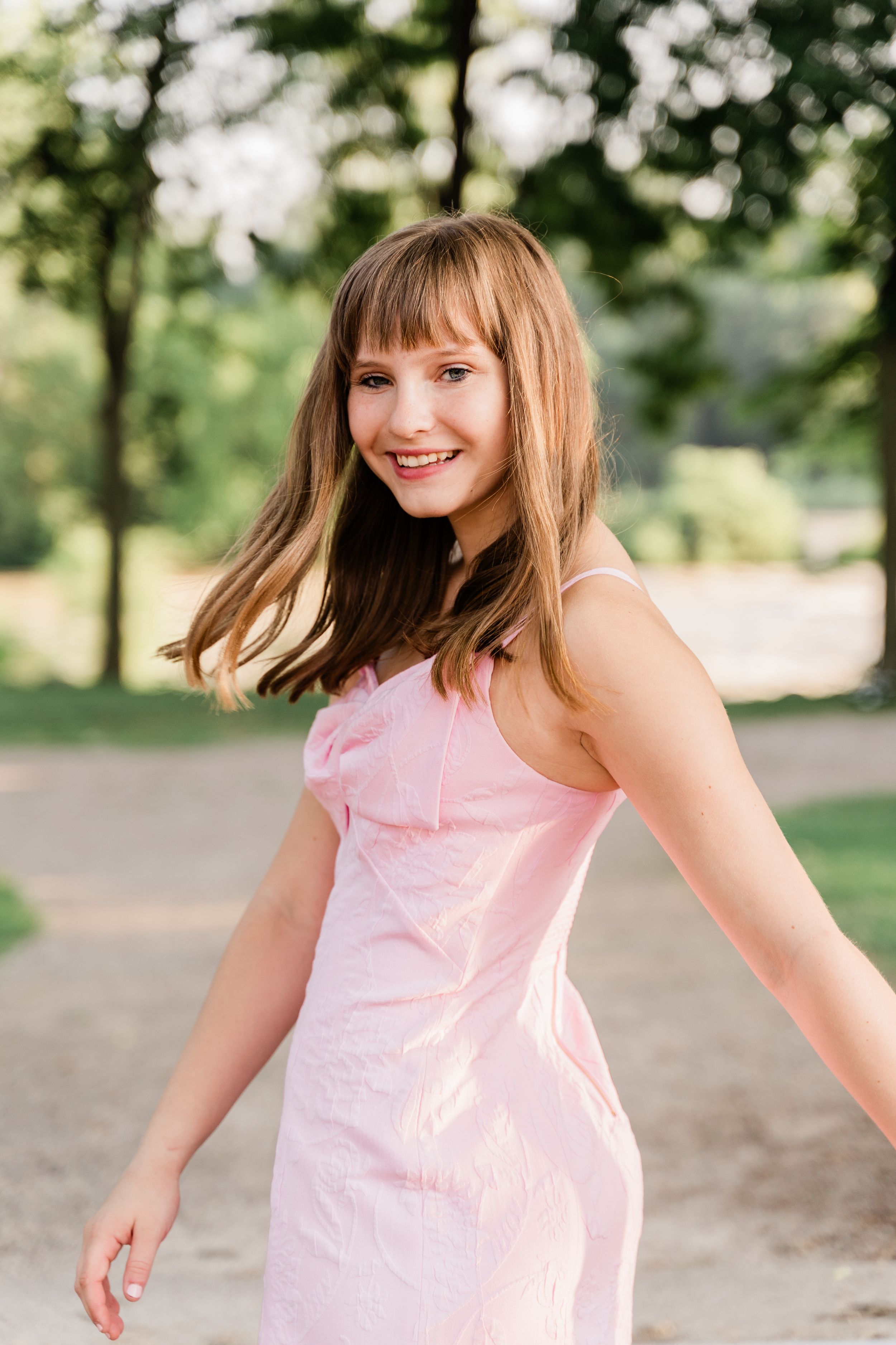 A person in a light pink dress twirls playfully outdoors in a park setting with trees in the background.