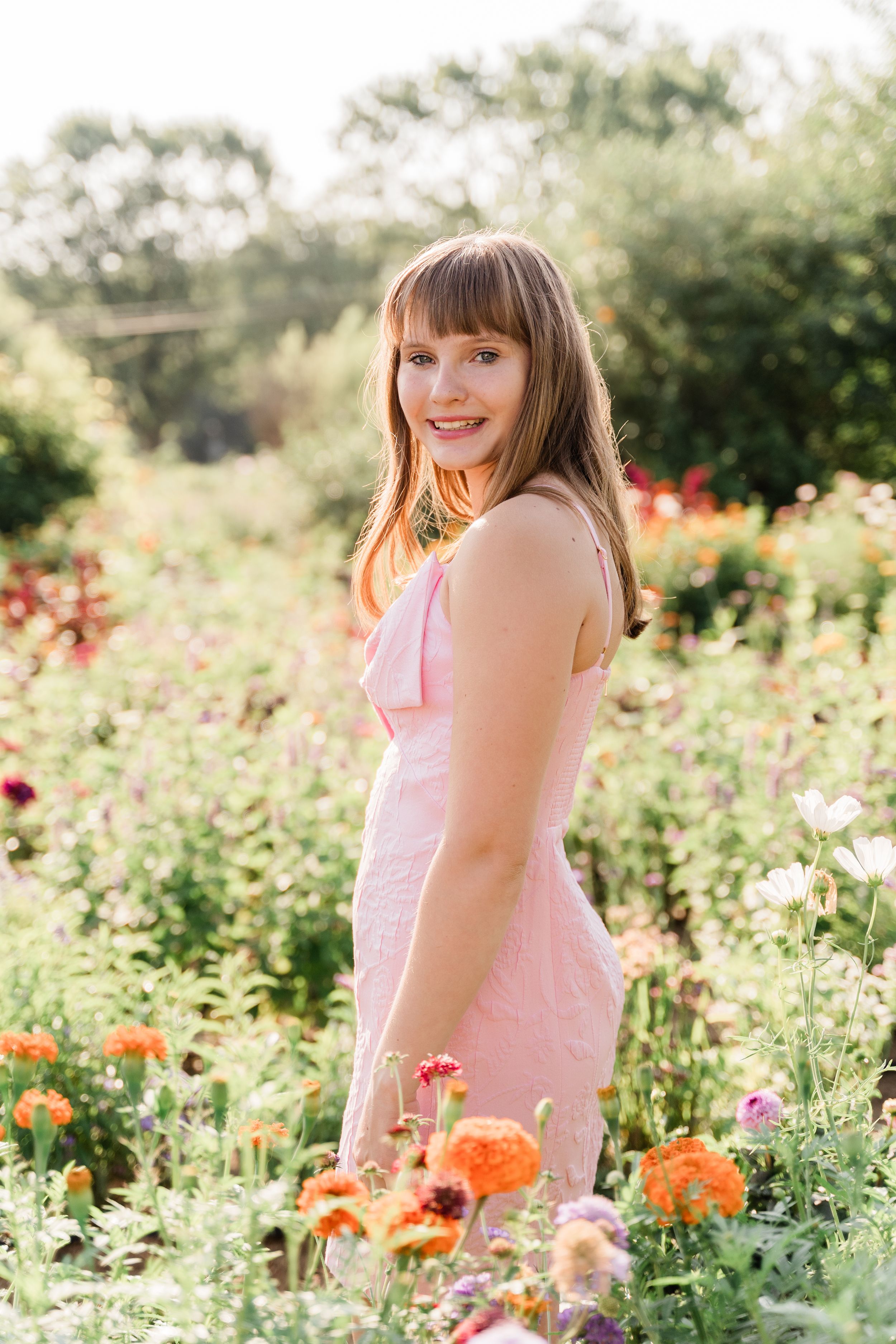 A person in a pink dress stands among vibrant orange and pink wildflowers in a sunny garden setting.