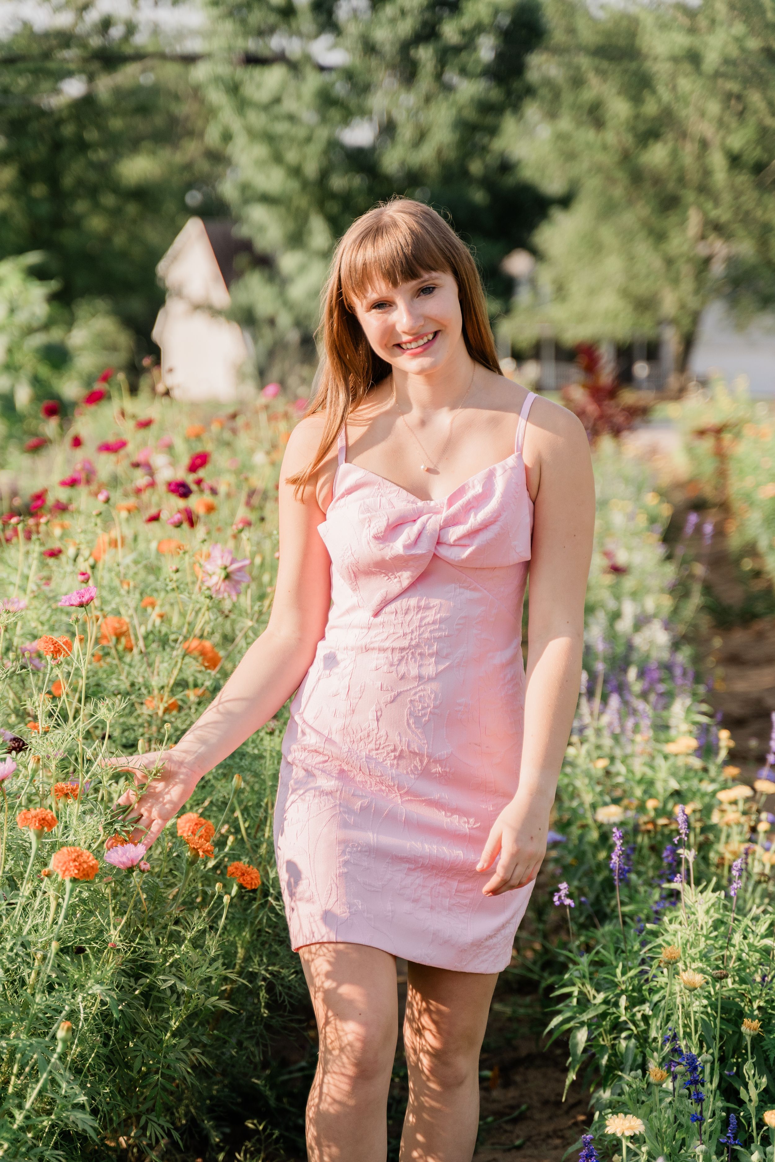 Woman in pink dress stands among colorful wildflowers and poppies in a sunny garden setting.
