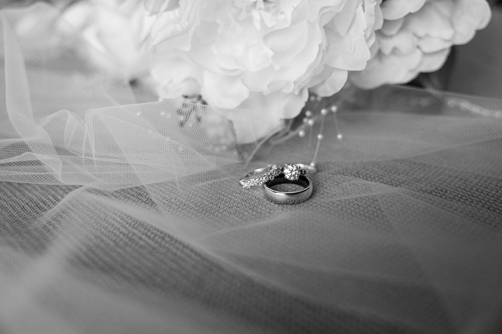 Black and white photo of wedding rings and diamond necklace resting on bridal veil with white floral bouquet.
