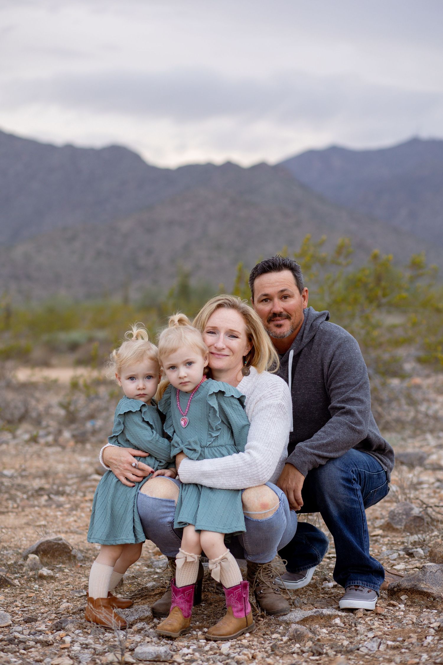 Family of four crouches together in rocky desert landscape with mountain backdrop at dusk.