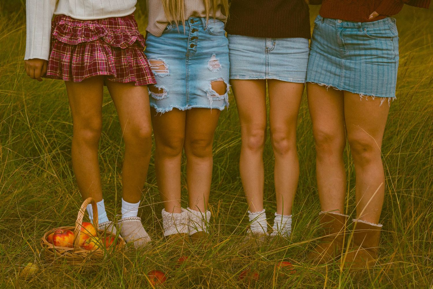 Four pairs of legs wearing skirts and sneakers stand in grass with a small wicker basket visible on the ground.