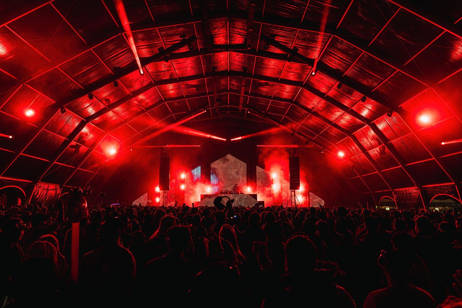 Concert venue illuminated in dramatic red lighting with silhouettes of crowd visible under a large arched ceiling structure.