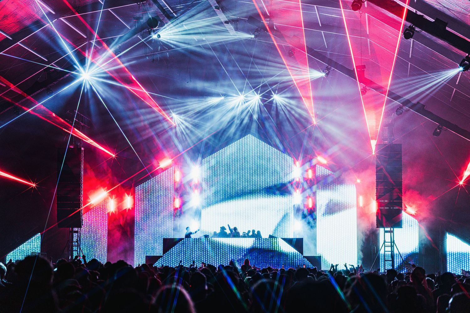 Colorful laser lights illuminate a concert venue with blue and pink beams cutting through atmospheric haze over a crowd.