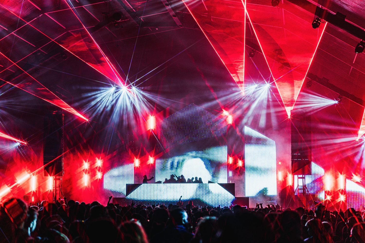 Concert crowd illuminated by dramatic red lighting and laser beams during a nighttime music festival performance.