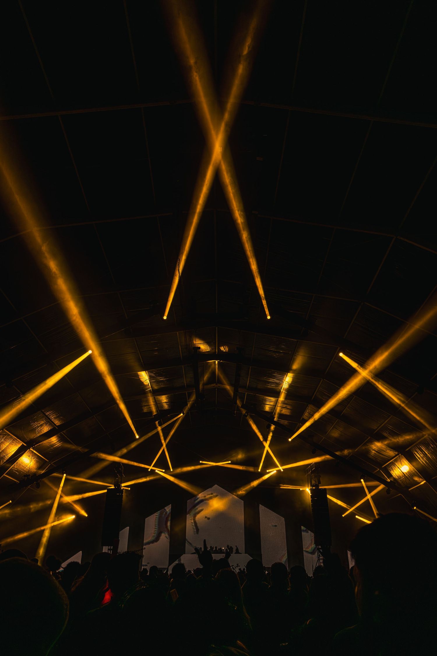 Dramatic golden spotlights create an X pattern on a dark ceiling at a nightclub or concert venue while crowd gathers below.