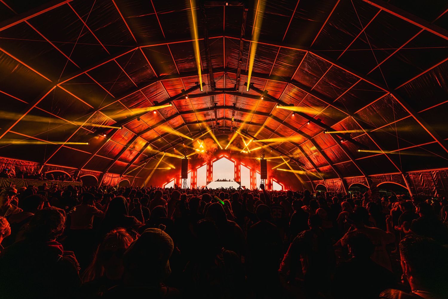 Concert crowd illuminated by vibrant orange and red stage lighting in a large indoor venue during a performance.