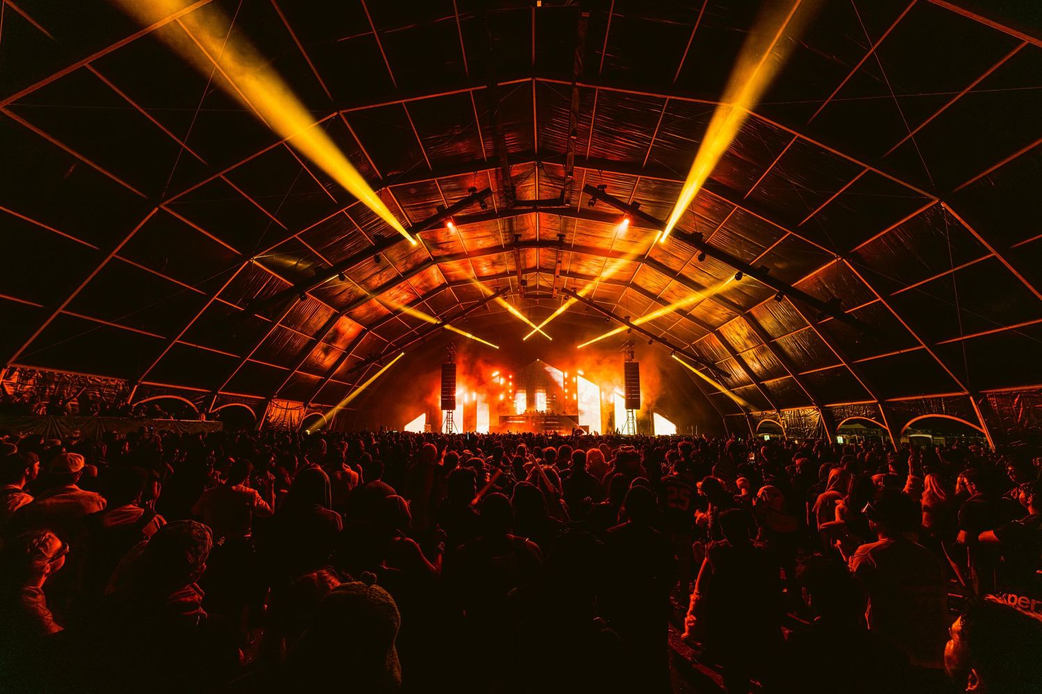 Concert crowd silhouetted against illuminated stage with dramatic orange lighting and geometric dome ceiling structure.