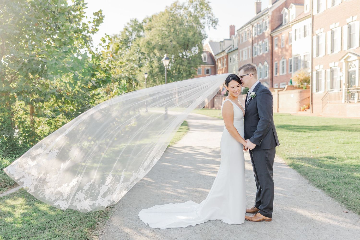 A couple shares a romantic moment on a sunlit path as the bride's veil flows dramatically in the breeze.