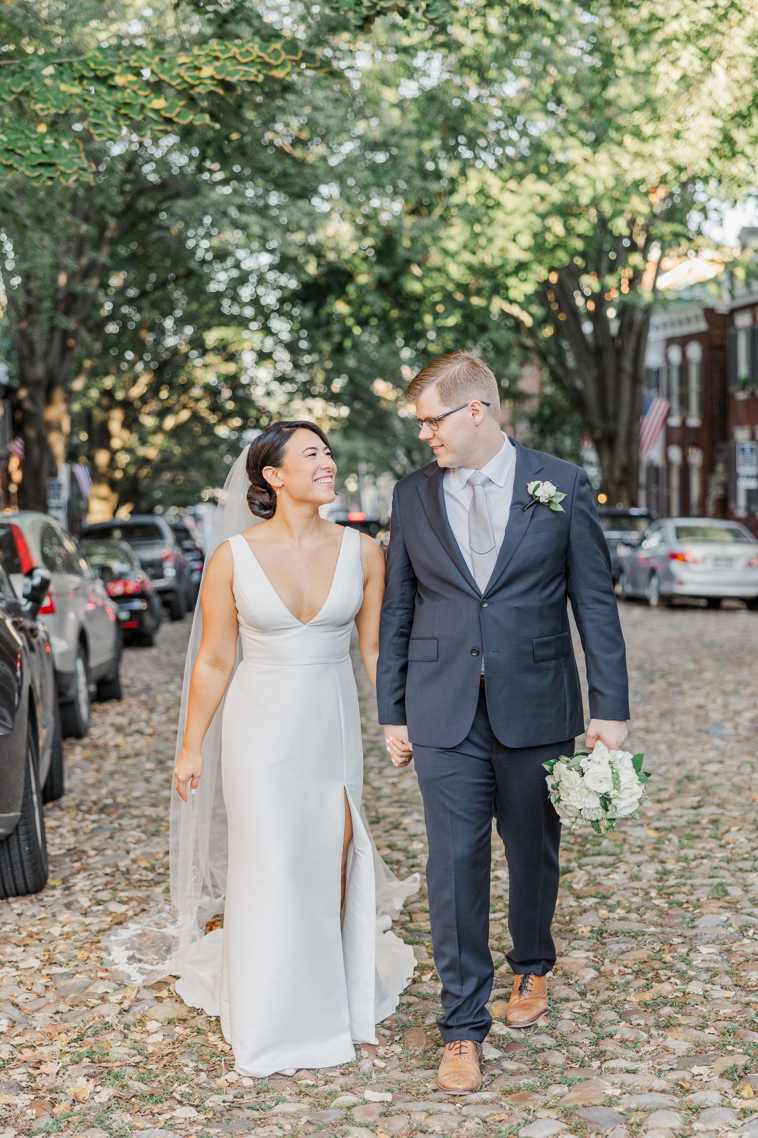 A bride and groom walk together on a cobblestone street lined with autumn trees while holding wine glasses.