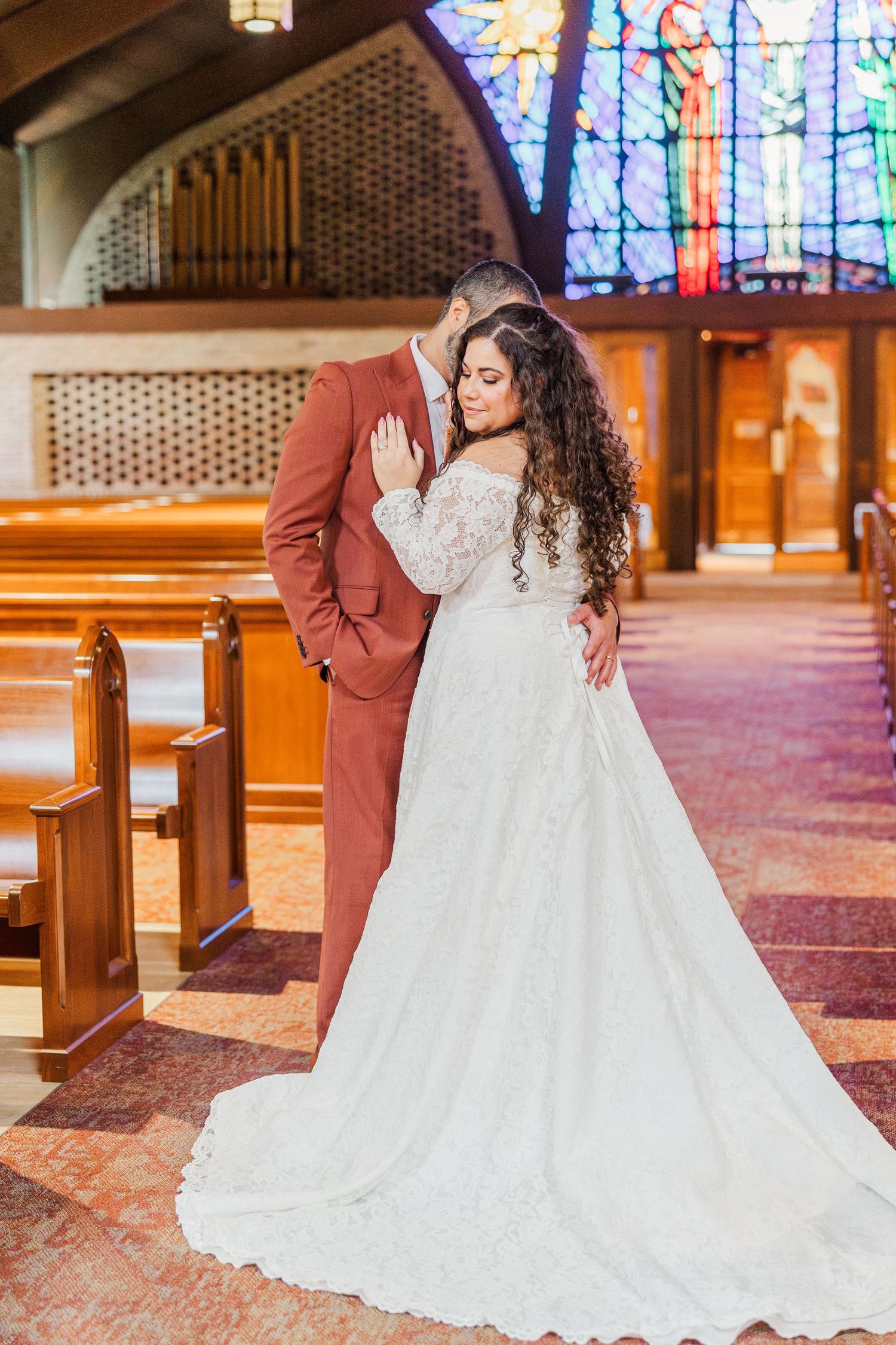 A bride in a flowing white lace wedding gown poses elegantly in a church with stained glass windows in the background.