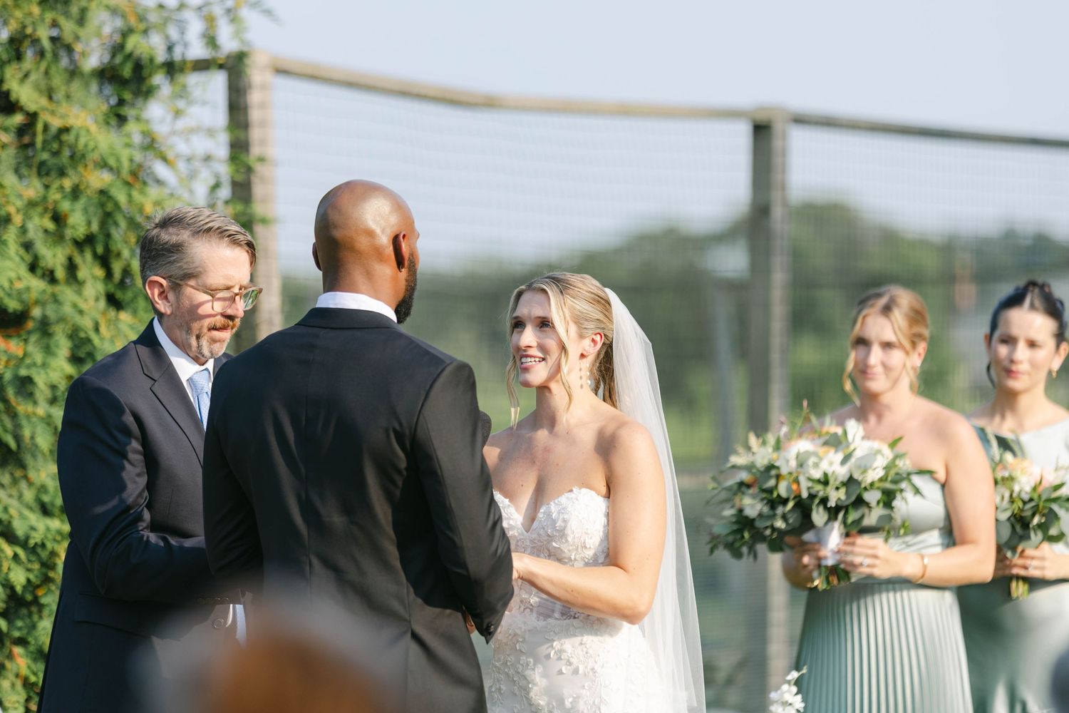 An outdoor wedding ceremony takes place on a sunny day with bridesmaids holding white floral bouquets. - Elawa Farm