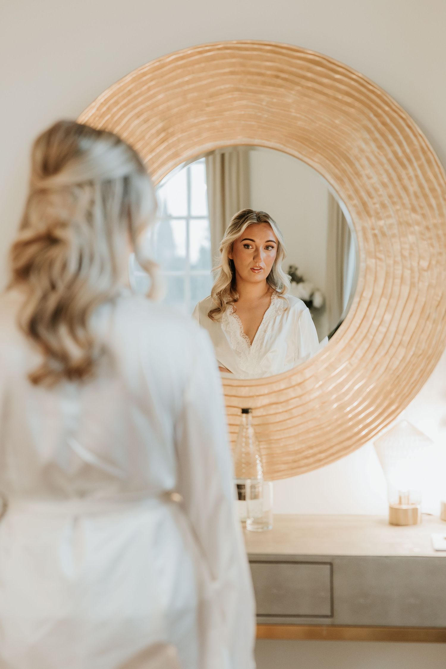 Reflection in a round woven mirror shows someone wearing a white robe in a bright, modern bathroom interior.
