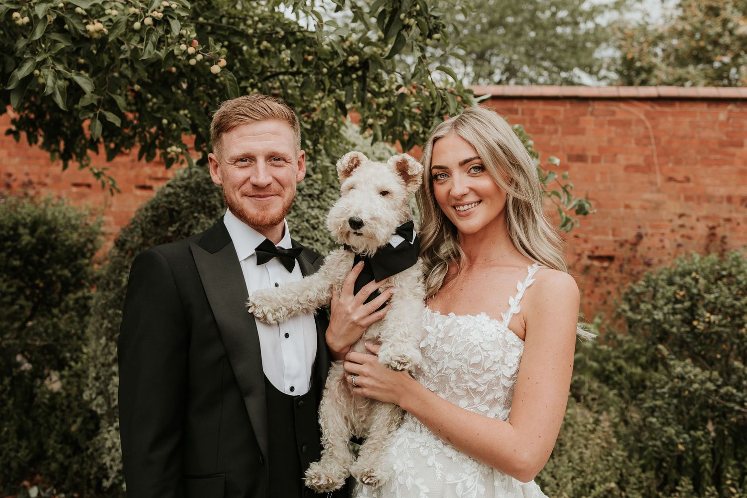 A couple in wedding attire poses with their white dog in a garden setting with brick walls and greenery.