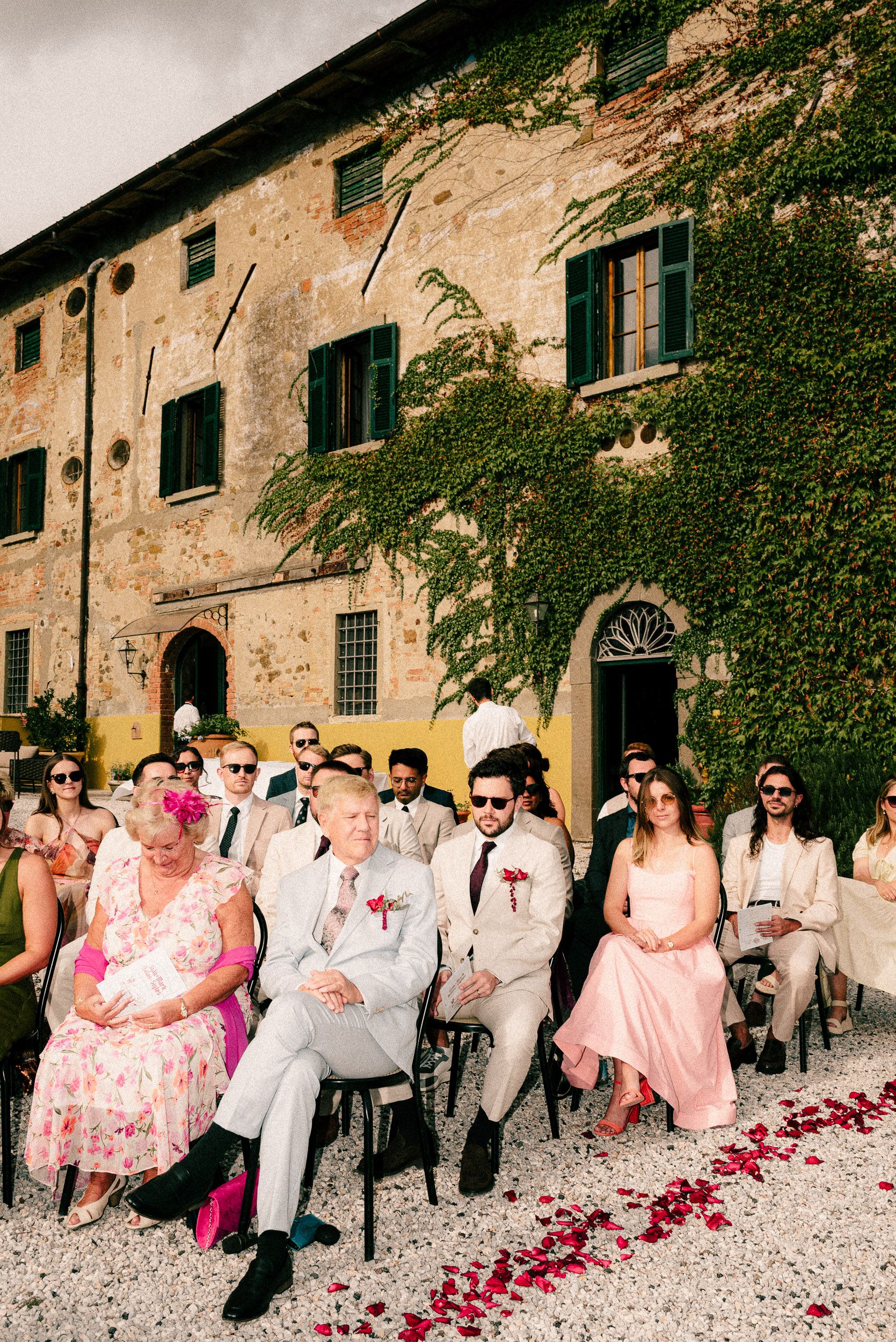 Wedding guests seated outdoors at a rustic Italian villa with ivy-covered walls and rose petals lining the aisle.