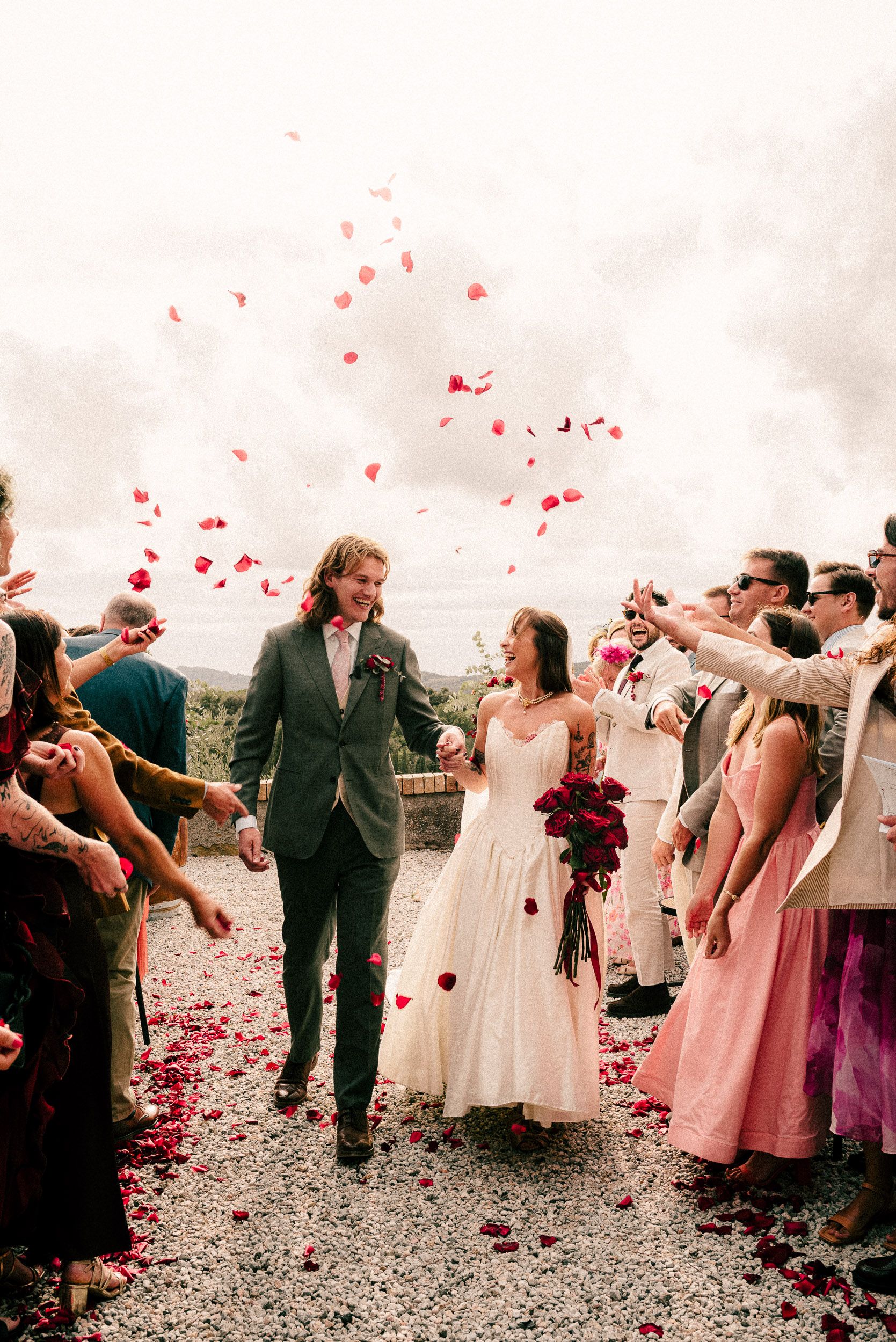 Wedding guests shower couple with red rose petals as they walk down gravel path during outdoor celebration.