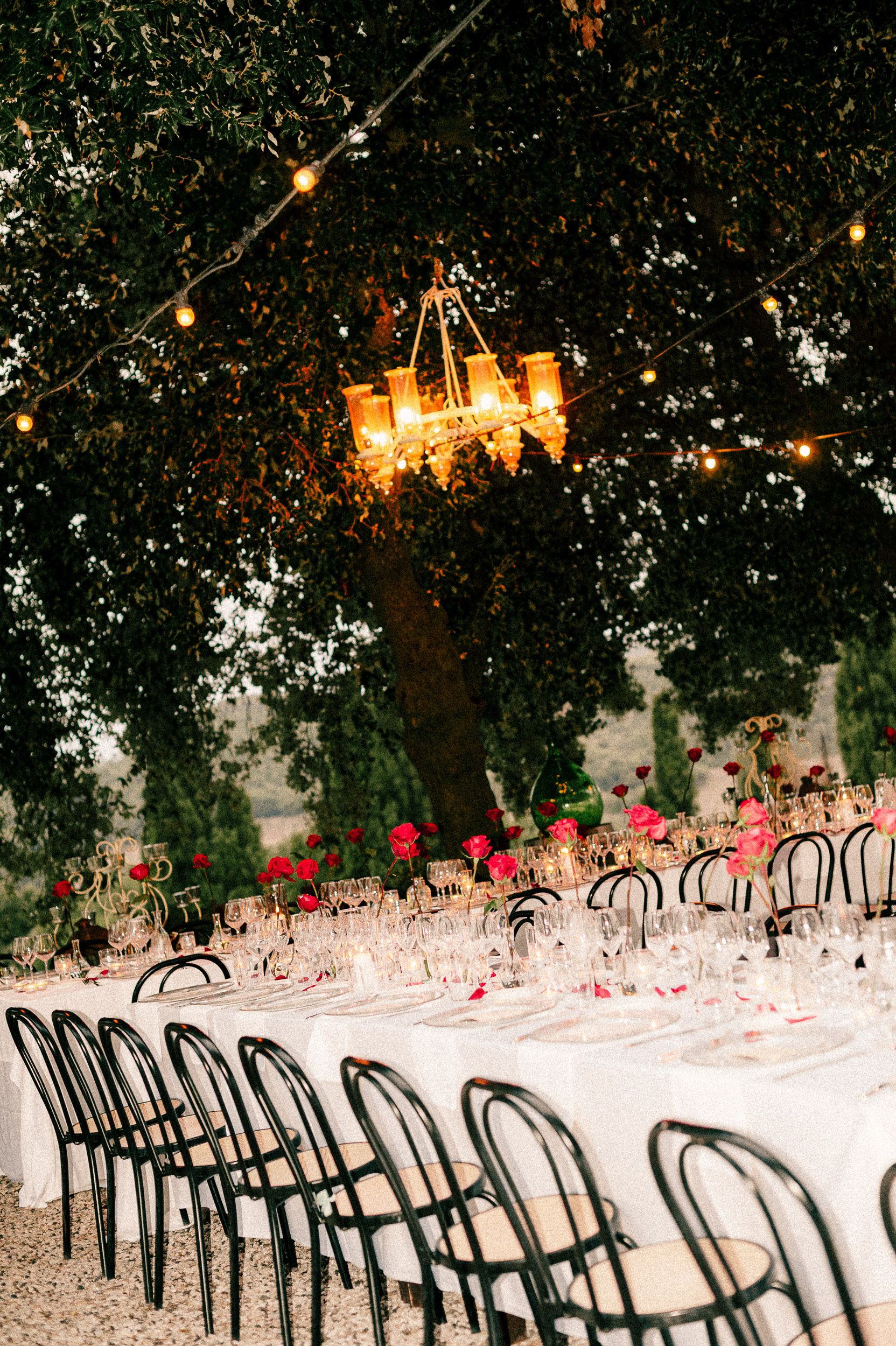 Elegant outdoor dining setup with black bentwood chairs and white tablecloths under string lights and tree canopy.
