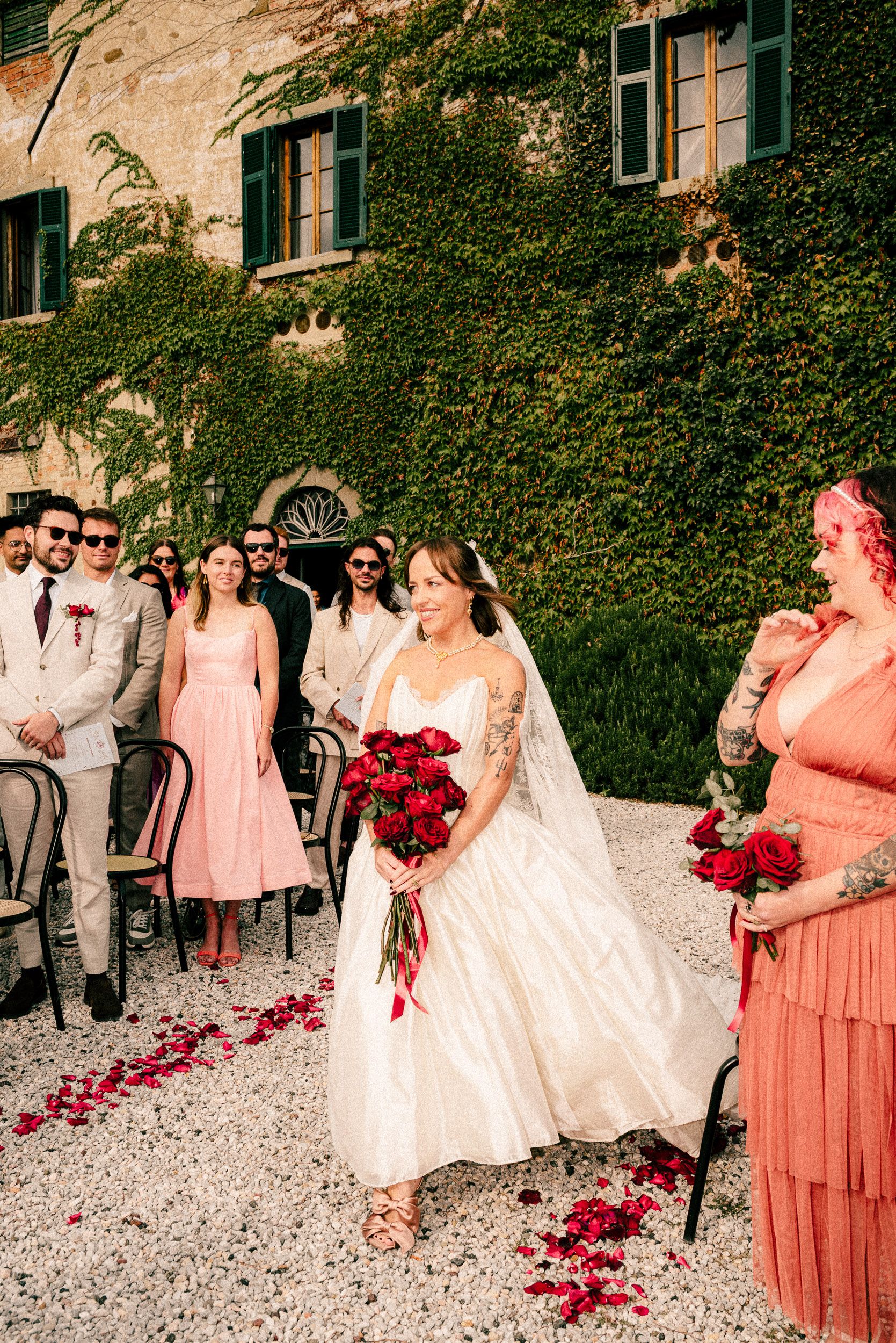 Wedding party members walk along a gravel path with scattered red rose petals and ivy-covered walls in the background.