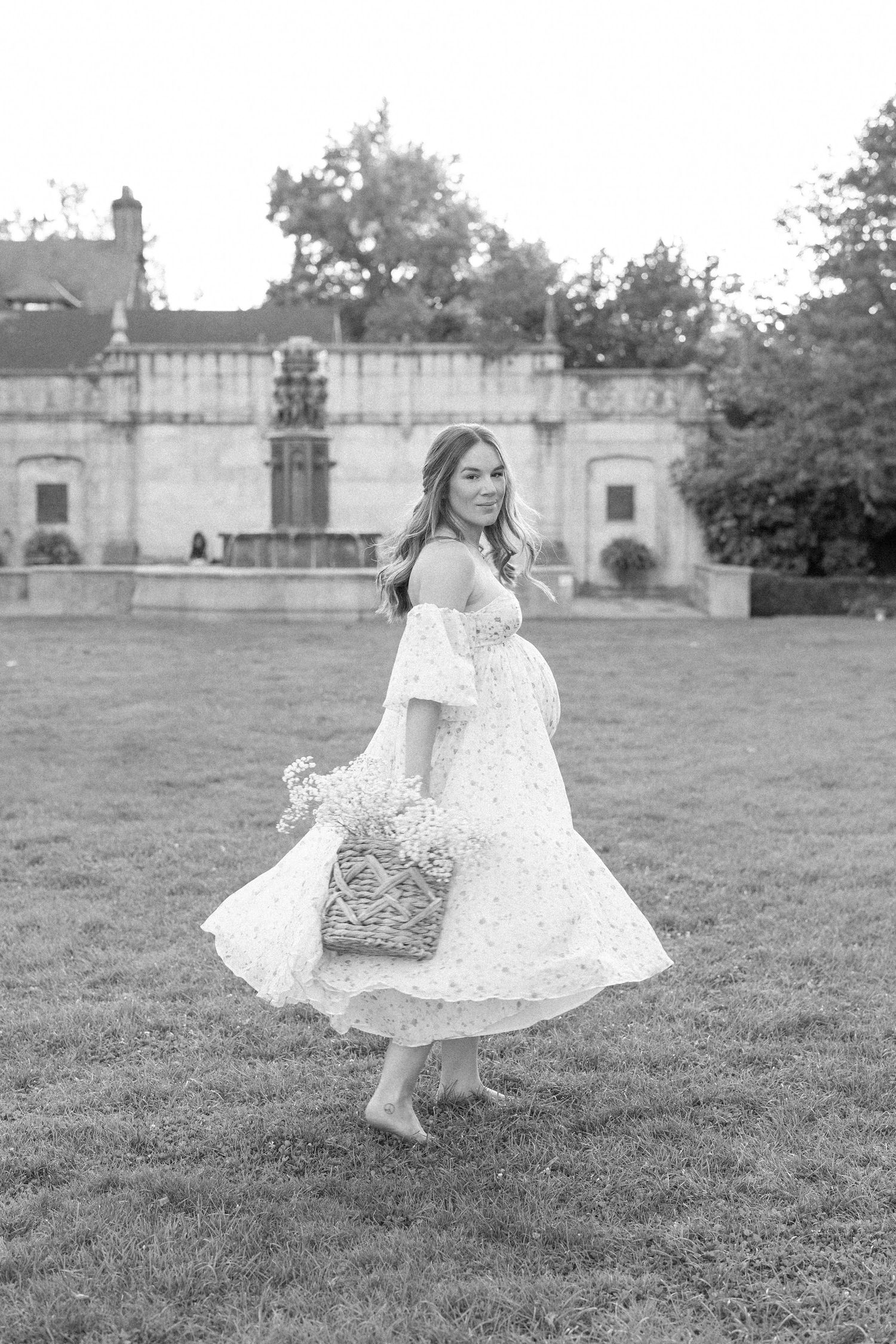 A woman in a flowing white lace dress twirls on the lawn of a historic estate in black and white photography.