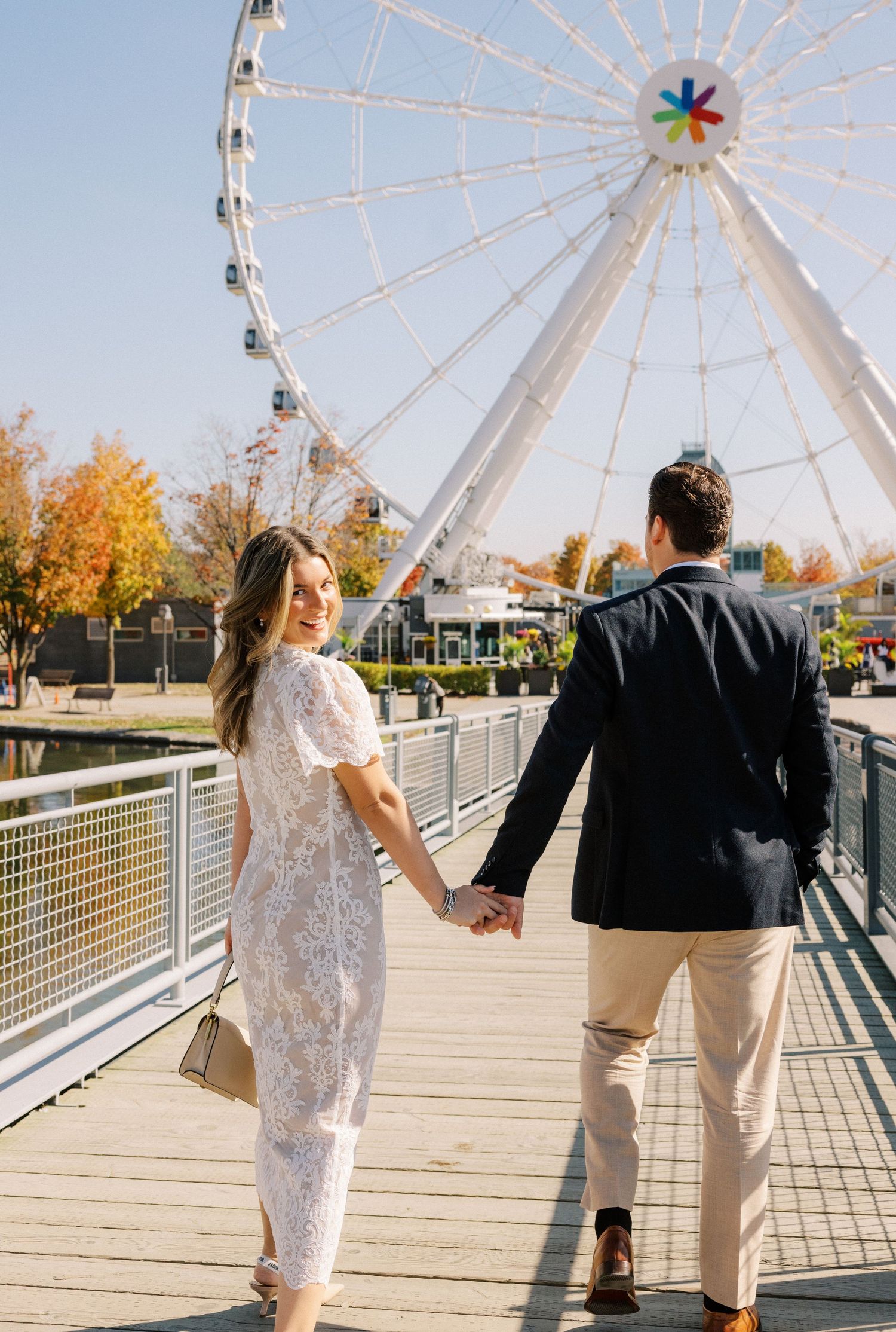 A couple holds hands while walking on a boardwalk with a colorful ferris wheel in the background.