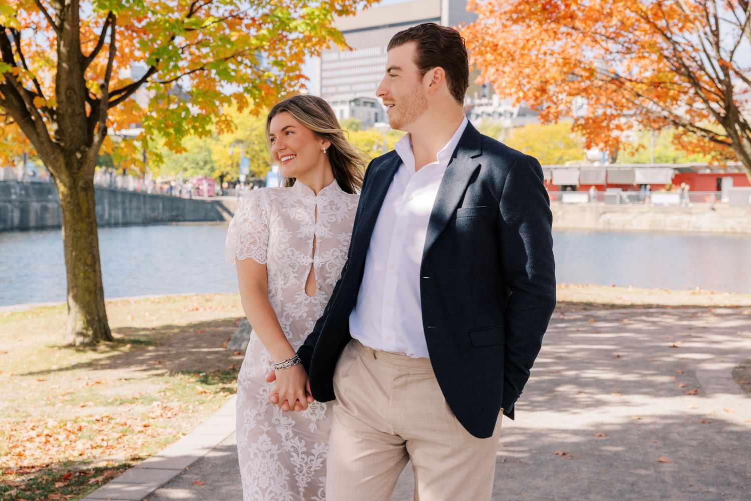 A couple in wedding attire walks together beneath vibrant orange and yellow autumn trees in a park setting.