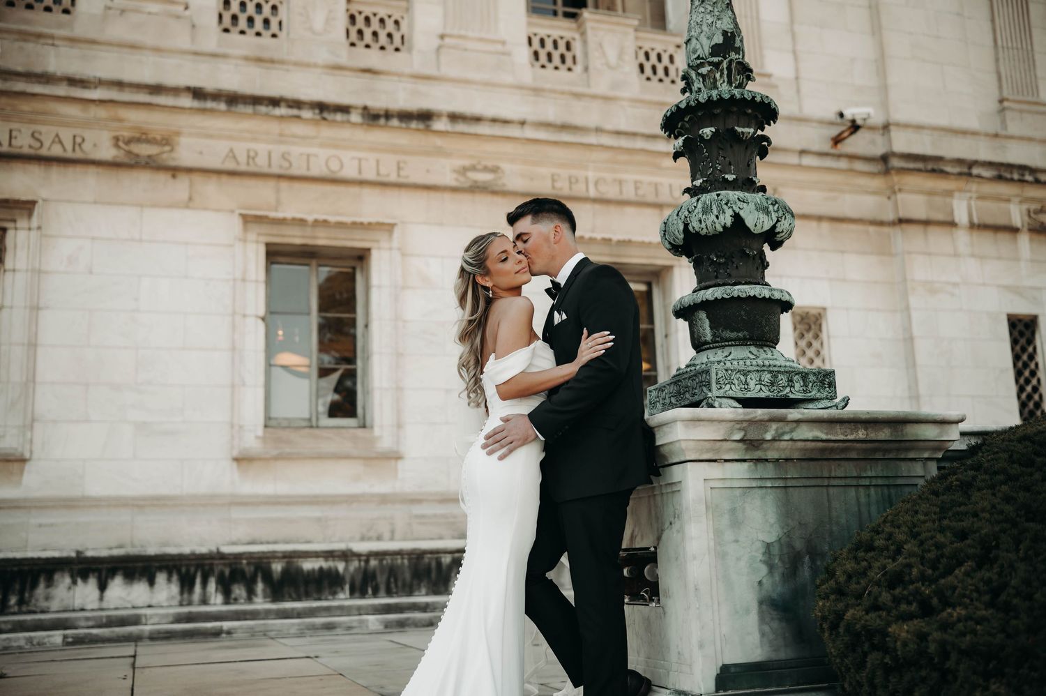 A bride in a fitted white gown and groom in a black suit embrace near an ornate fountain.