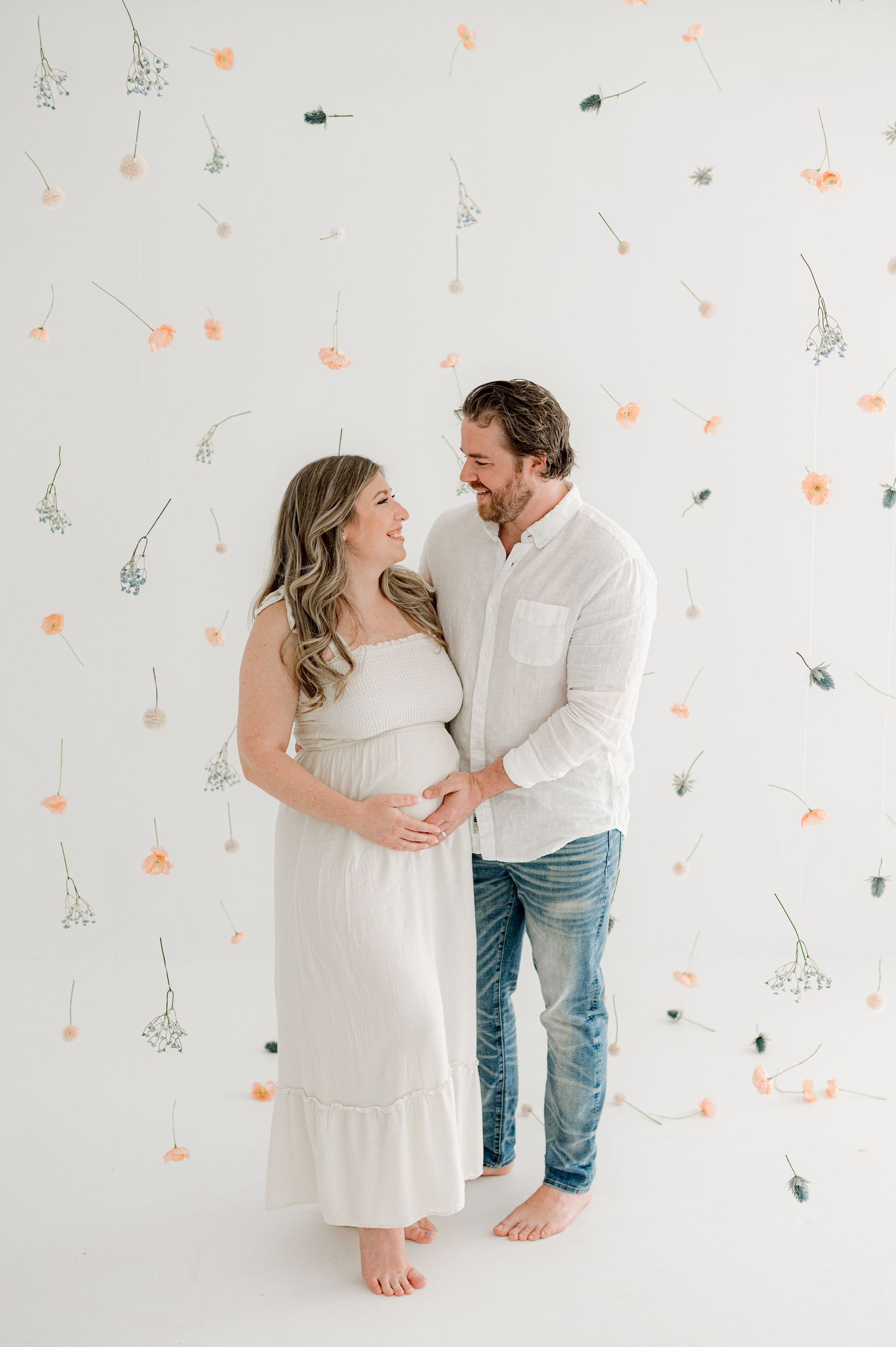 A couple shares joyful moments against a light backdrop with delicate floral wall decorations at maternity photoshoot Jacksonville session.