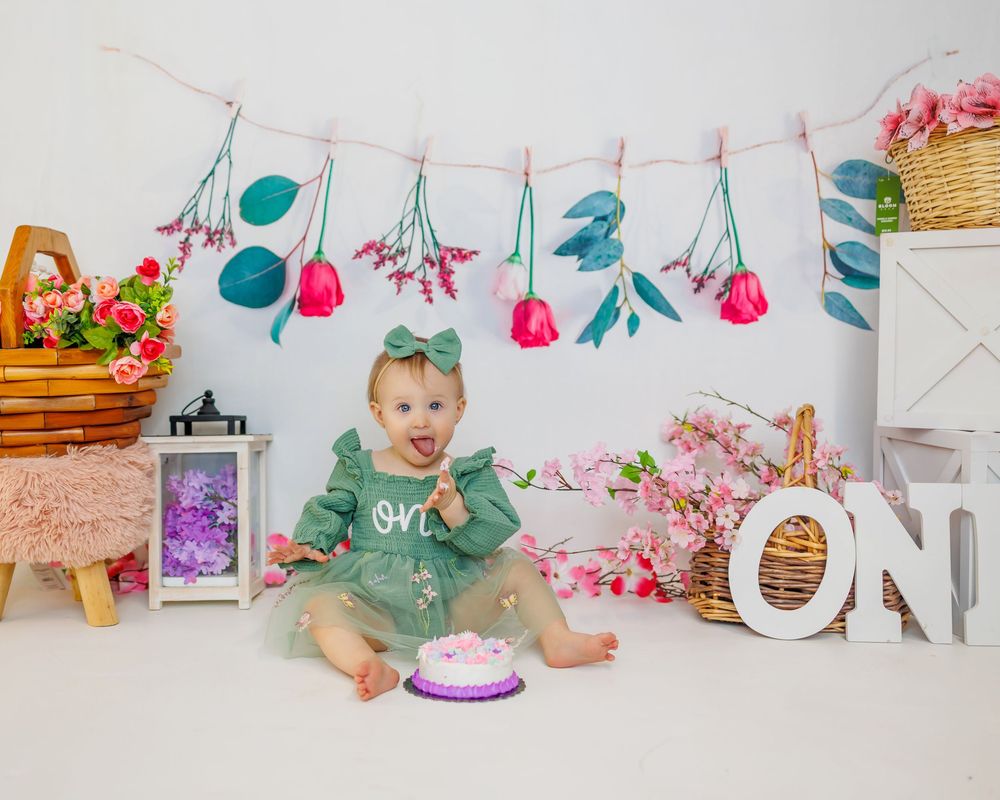 A baby in a green outfit sits on a white backdrop with pink and teal party decorations and a cake for a first birthday.