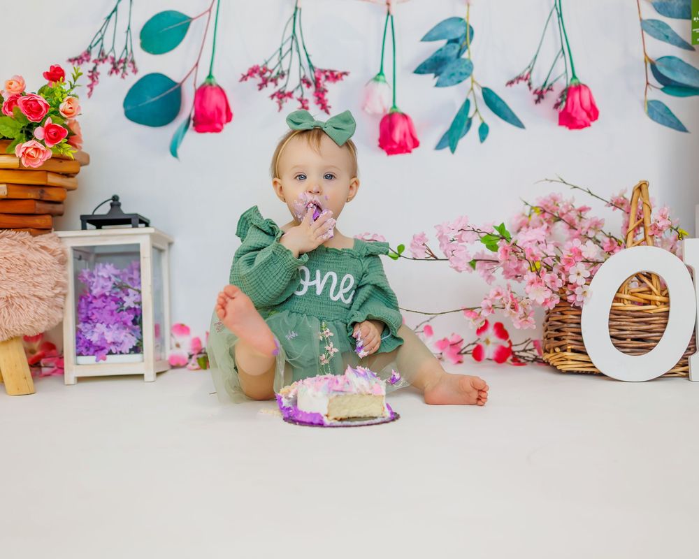 A baby in a green shirt sits among pink flowers and white decorations during a cake smash photo session.