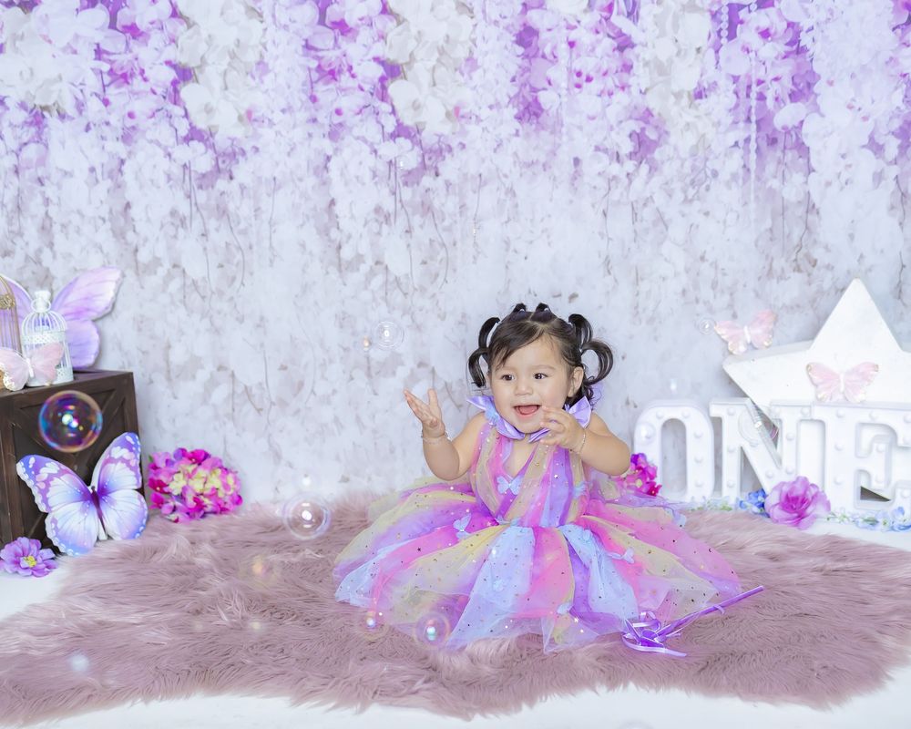 A child in a pink tutu dress sits on a rustic backdrop with purple flower petals scattered around.