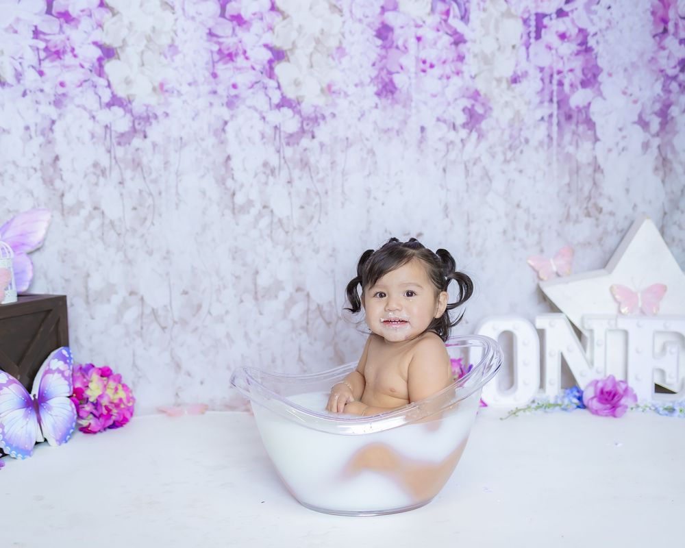 A baby relaxes in a white bathtub surrounded by purple floral decorations and white bunny props during a photo session.