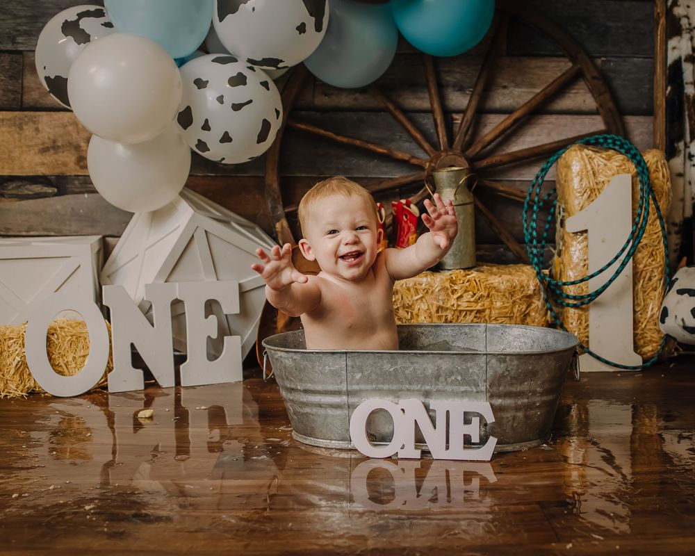 First birthday photo setup with rustic elements, balloons, and vintage metal washtub on wooden floor.