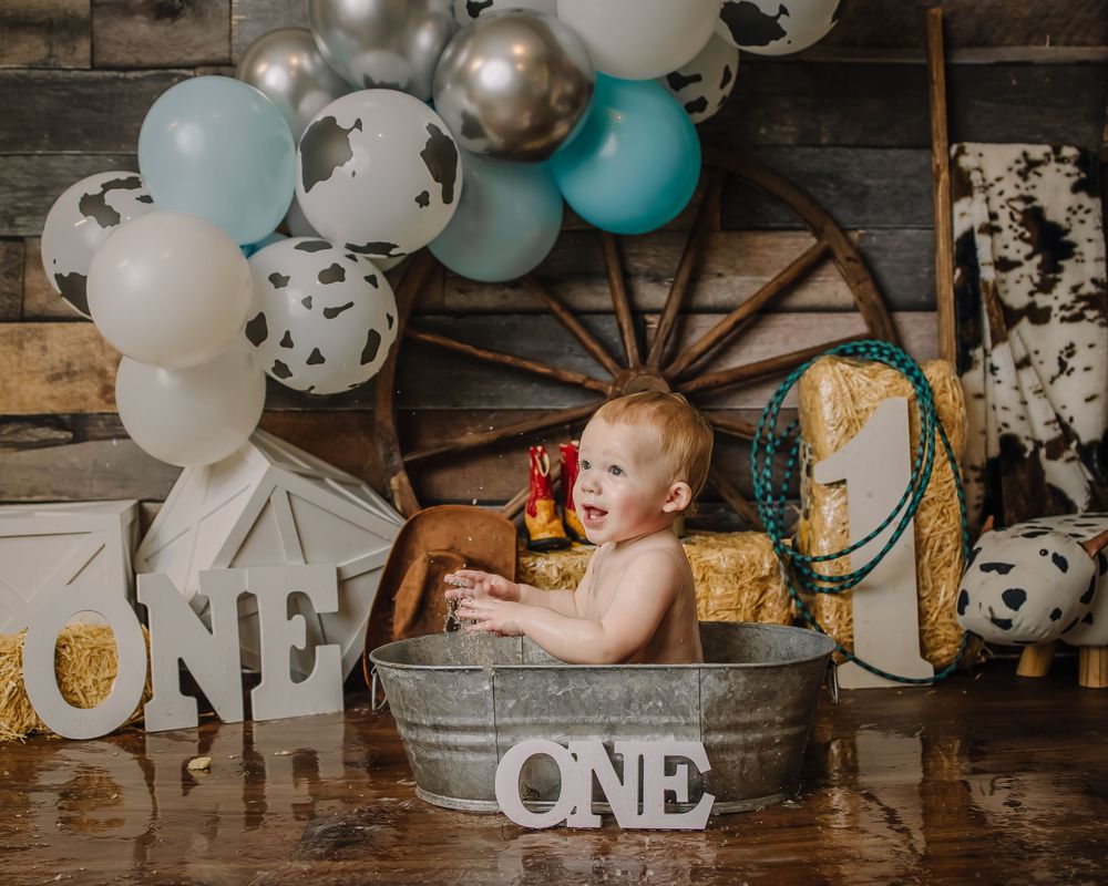 Rustic first birthday party setup with white and blue balloons, wooden accents, and 'ONE' lettering against barn wood wall.