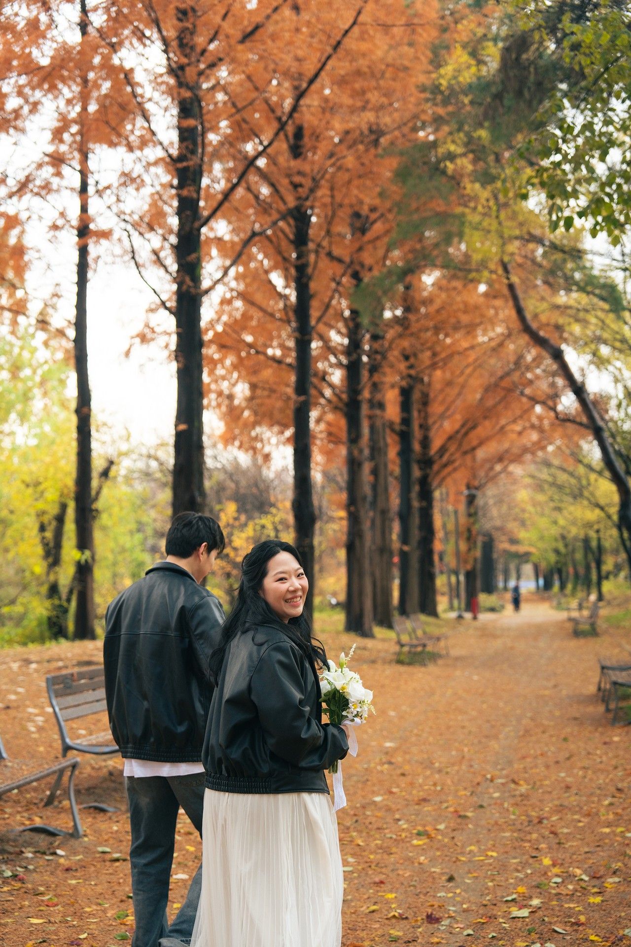 A couple walks along a tree-lined autumn path with orange foliage and fallen leaves creating a romantic scene.