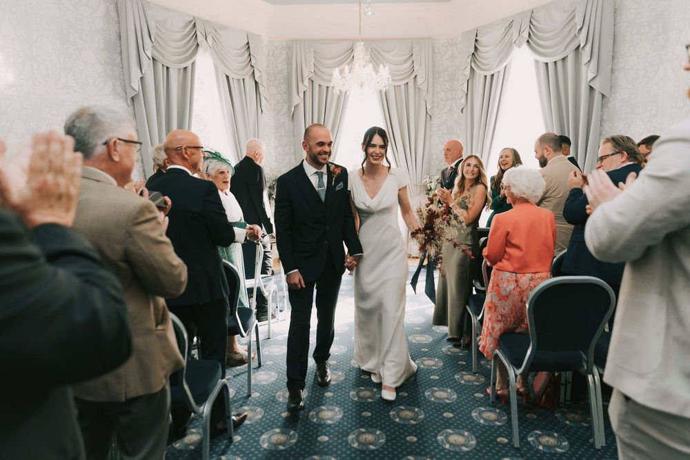 Wedding guests gather and mingle during a reception in an elegant ballroom with white curtains and classic decor.