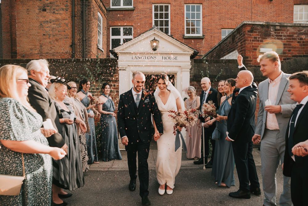Wedding guests line up along a red brick building exterior to celebrate during a daytime reception.
