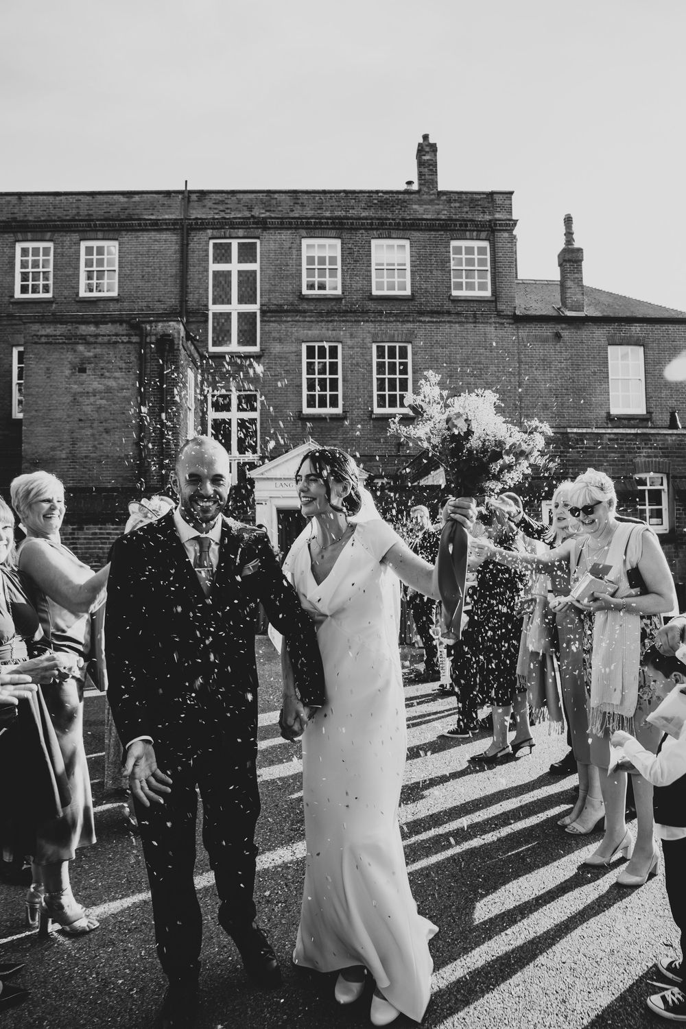 Wedding guests throw confetti at the newlyweds during an outdoor celebration on a brick courtyard.