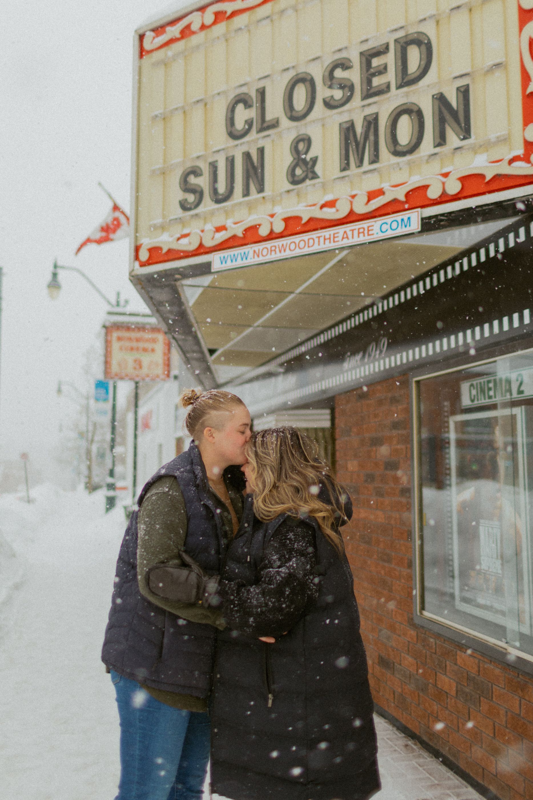 A romantic moment outside a theater with 'Closed Sun & Mon' sign during snowfall.