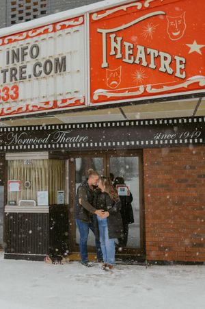Two people embrace outside a vintage theatre entrance during a snowstorm.