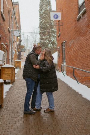A couple shares a romantic moment on a brick pathway between buildings during snowfall.